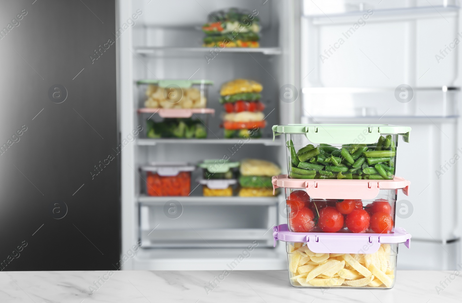 Photo of Containers with different frozen vegetables on white marble table near open refrigerator