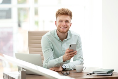 Photo of Portrait of handsome young man with smartphone at desk in office