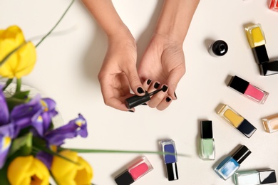 Woman applying nail polish near bottles on white table with flowers, top view