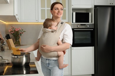 Mother holding her child in sling (baby carrier) while cooking pasta in kitchen