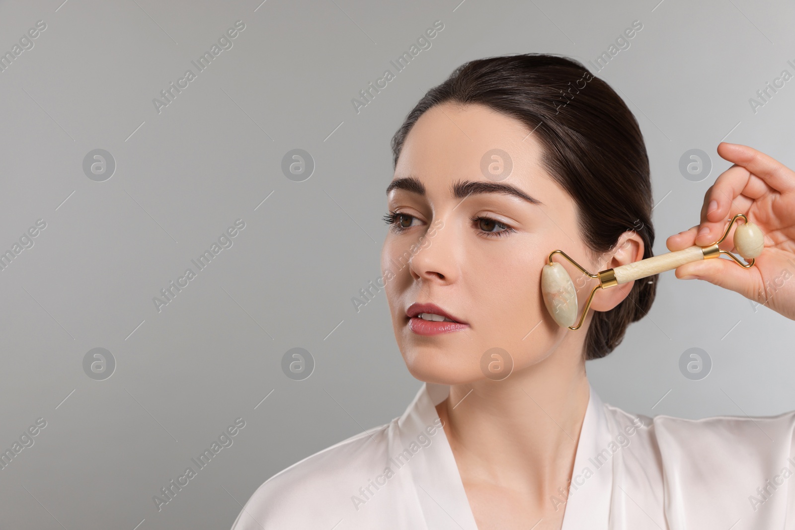 Photo of Young woman massaging her face with jade roller on grey background. Space for text