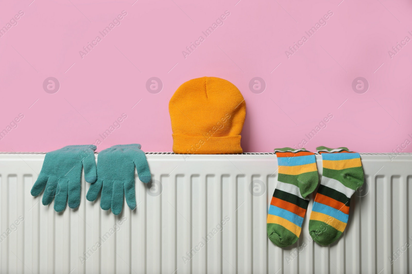 Photo of Modern radiator with knitted hat, socks and gloves near pink wall indoors