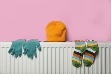 Photo of Modern radiator with knitted hat, socks and gloves near pink wall indoors