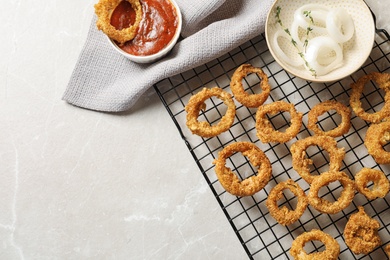 Cooling rack with homemade crunchy fried onion rings and sauces on light background, top view. Space for text