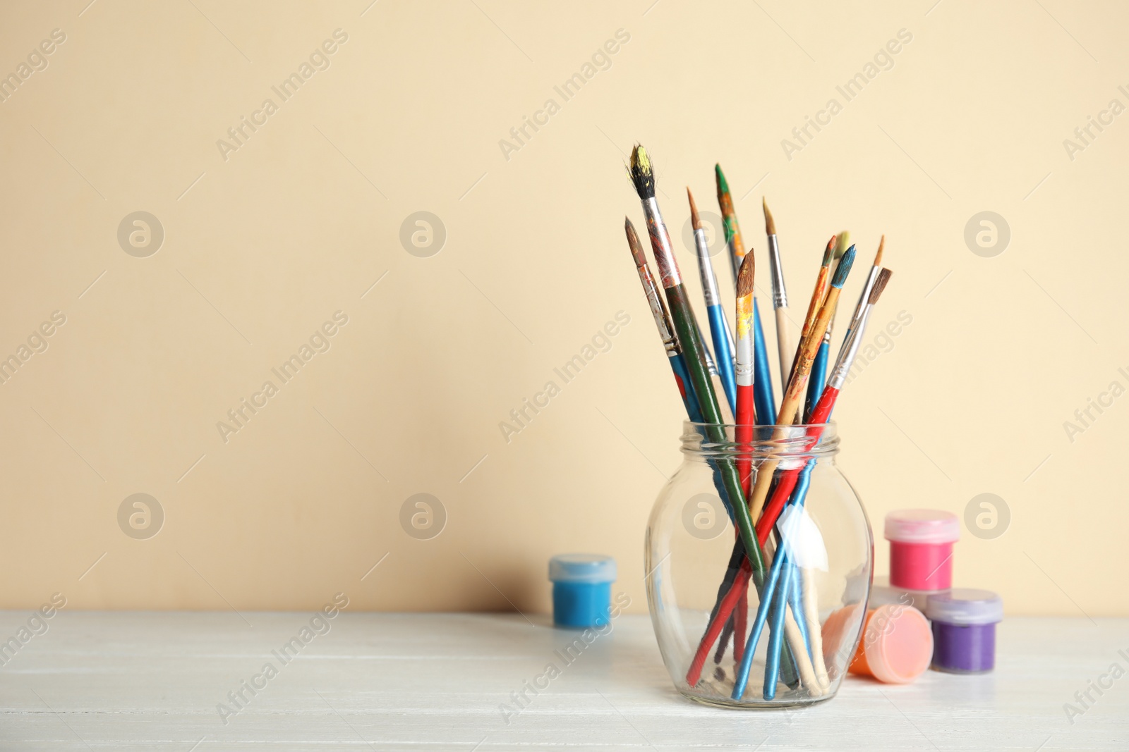 Photo of Glass with brushes and paint jars on table against color background. Space for text
