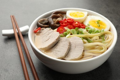 Photo of Delicious ramen with meat in bowl and chopsticks on grey textured table, closeup. Noodle soup