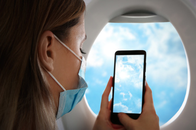 Image of Traveling by airplane during coronavirus pandemic. Woman with face mask taking photo of sky through porthole