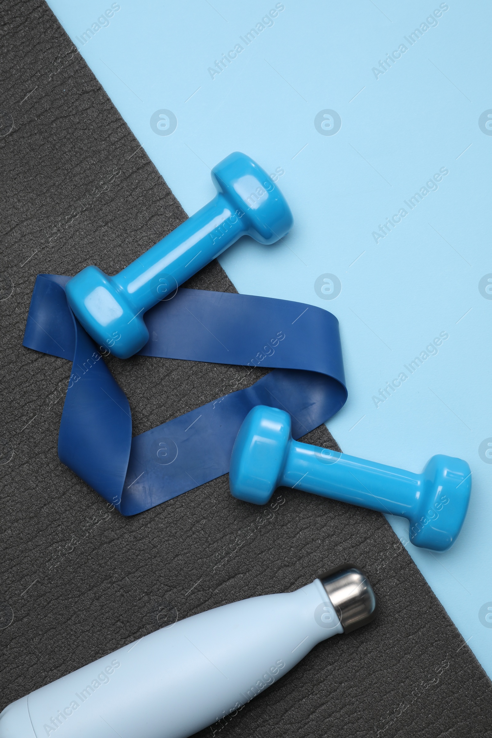 Photo of Two dumbbells, yoga mat, fitness elastic band and thermo bottle on light blue background, flat lay