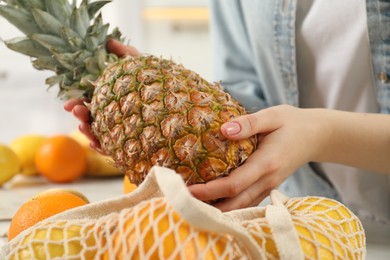 Photo of Woman taking pineapple out from string bag at table, closeup