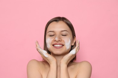 Photo of Young woman washing face with cleansing foam on pink background. Skin care cosmetic