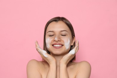 Young woman washing face with cleansing foam on pink background. Skin care cosmetic