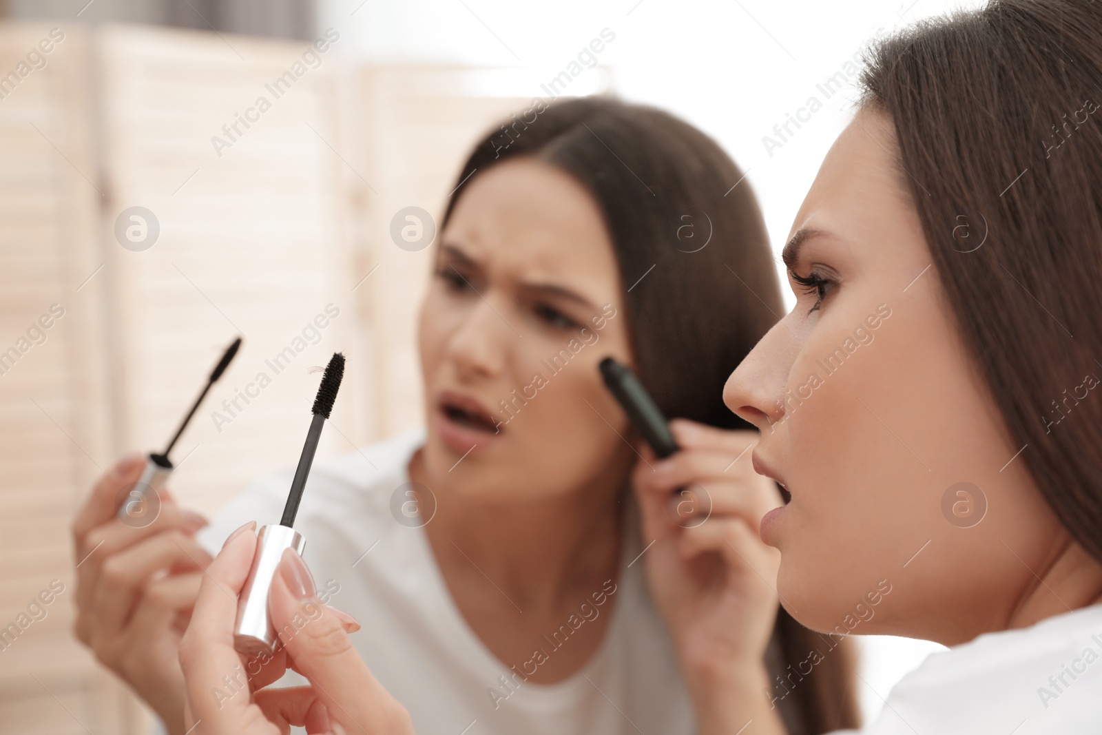 Photo of Beautiful woman holding mascara brush with fallen eyelashes near mirror indoors
