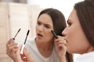Photo of Beautiful woman holding mascara brush with fallen eyelashes near mirror indoors