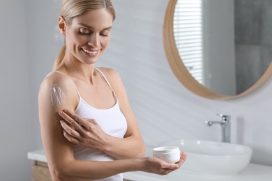 Photo of Happy woman applying body cream onto arm in bathroom, space for text