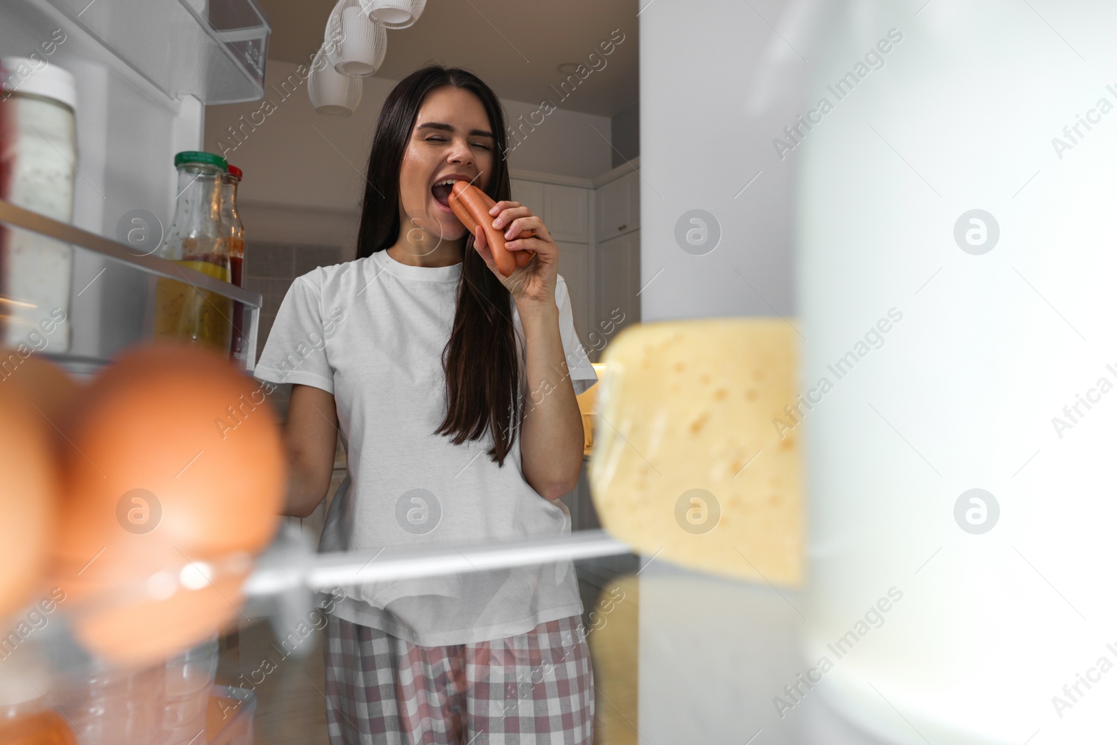 Photo of Young woman eating sausages near modern refrigerator in kitchen at night, view from inside