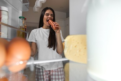 Young woman eating sausages near modern refrigerator in kitchen at night, view from inside