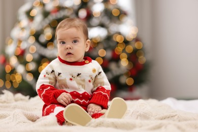 Photo of Cute little baby in Christmas sweater on knitted blanket against blurred festive lights, space for text. Winter holiday