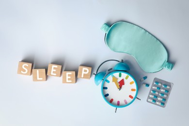 Photo of Word Sleep made of wooden cubes near blindfold, pills and alarm clock on white background, flat lay. Insomnia treatment
