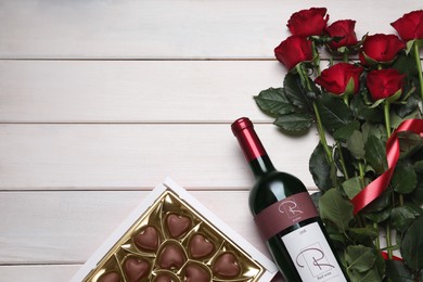 Bottle of red wine, beautiful roses and heart shaped chocolate candies on white wooden table, flat lay. Space for text