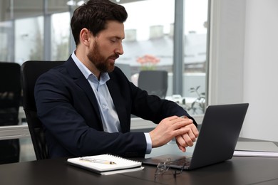 Man checking time on his watch while working with laptop at black desk in office
