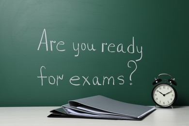 Photo of Office folders and alarm clock on white table near chalkboard with phrase Are You Ready For Exams