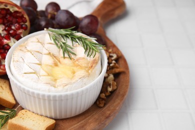 Photo of Board with tasty baked camembert and rosemary in bowl on white tiled table, closeup. Space for text