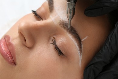 Beautician making permanent eyebrow makeup to young woman on white background, closeup