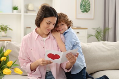 Photo of Little son congratulating his mom with Mother`s day at home. Woman holding handmade greeting card