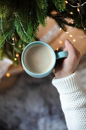 Photo of Woman holding cup of cocoa near Christmas tree indoors, top view