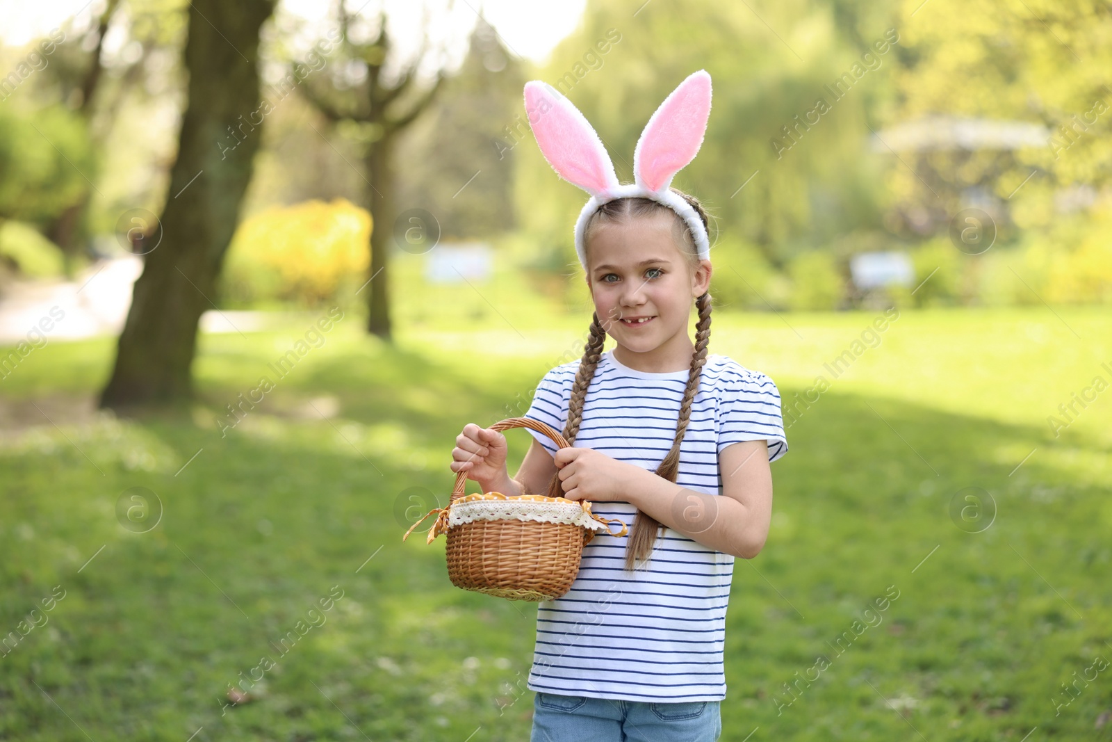 Photo of Easter celebration. Cute little girl in bunny ears holding wicker basket outdoors, space for text