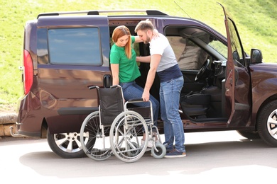 Photo of Young man helping woman to get out from van into wheelchair outdoors