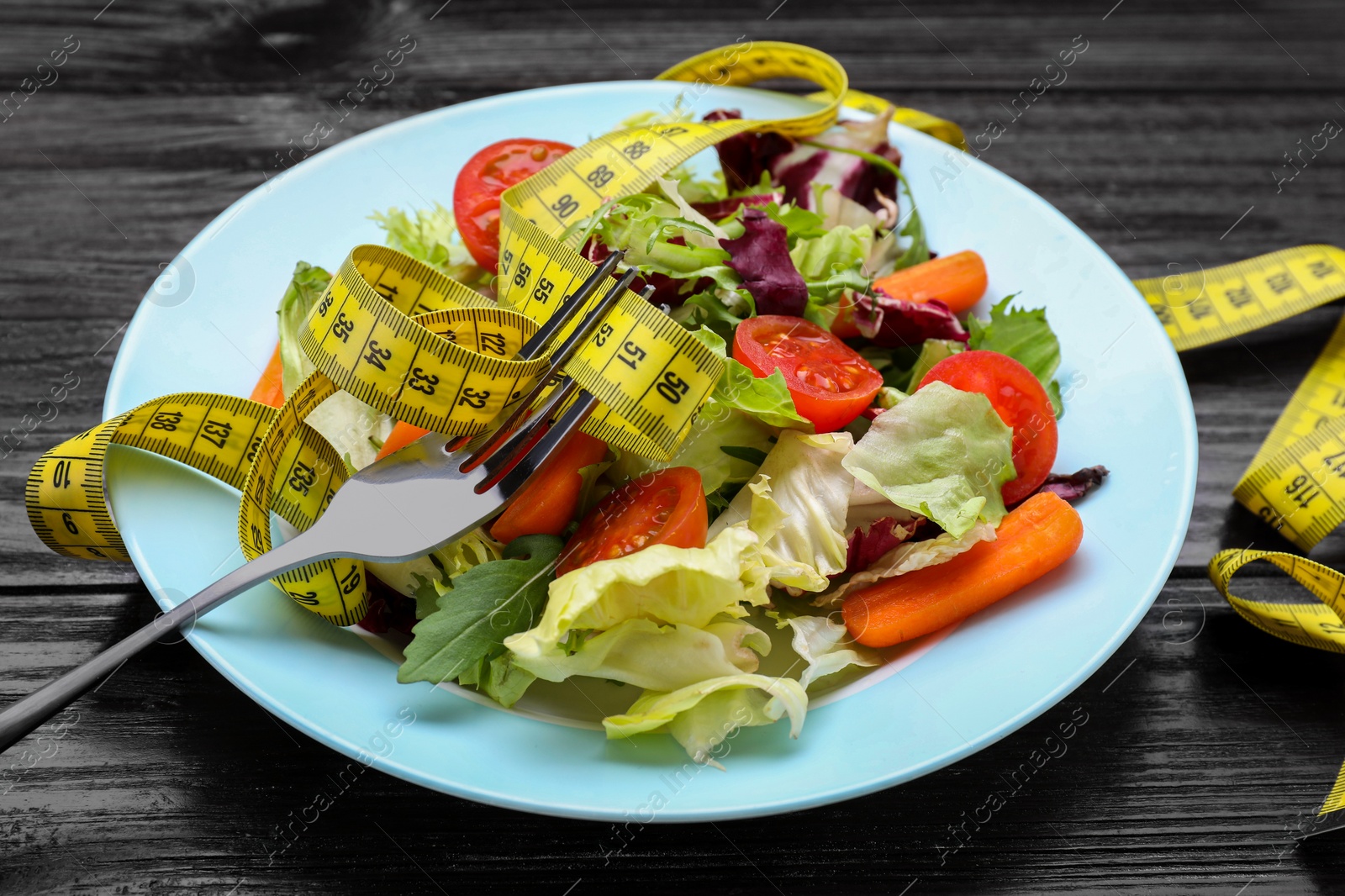 Photo of Plate with fresh vegetable salad, fork and measuring tape on wooden table, closeup. Healthy diet concept