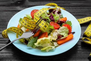 Photo of Plate with fresh vegetable salad, fork and measuring tape on wooden table, closeup. Healthy diet concept