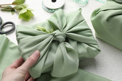Photo of Furoshiki technique. Woman holding gift packed in green fabric and decorated with hellebore flower at white wooden table, closeup
