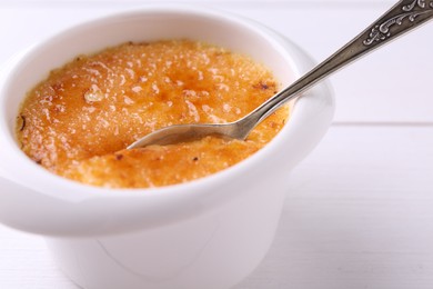 Photo of Taking delicious creme brulee with spoon from bowl at white wooden table, closeup