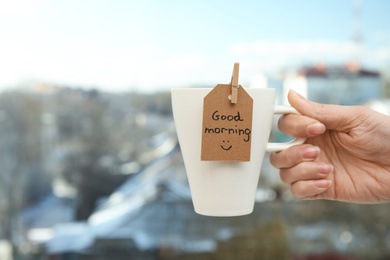 Photo of Woman holding cup of delicious coffee with GOOD MORNING wish near window