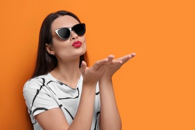 Photo of Beautiful young woman in stylish sunglasses blowing kiss on orange background. Space for text