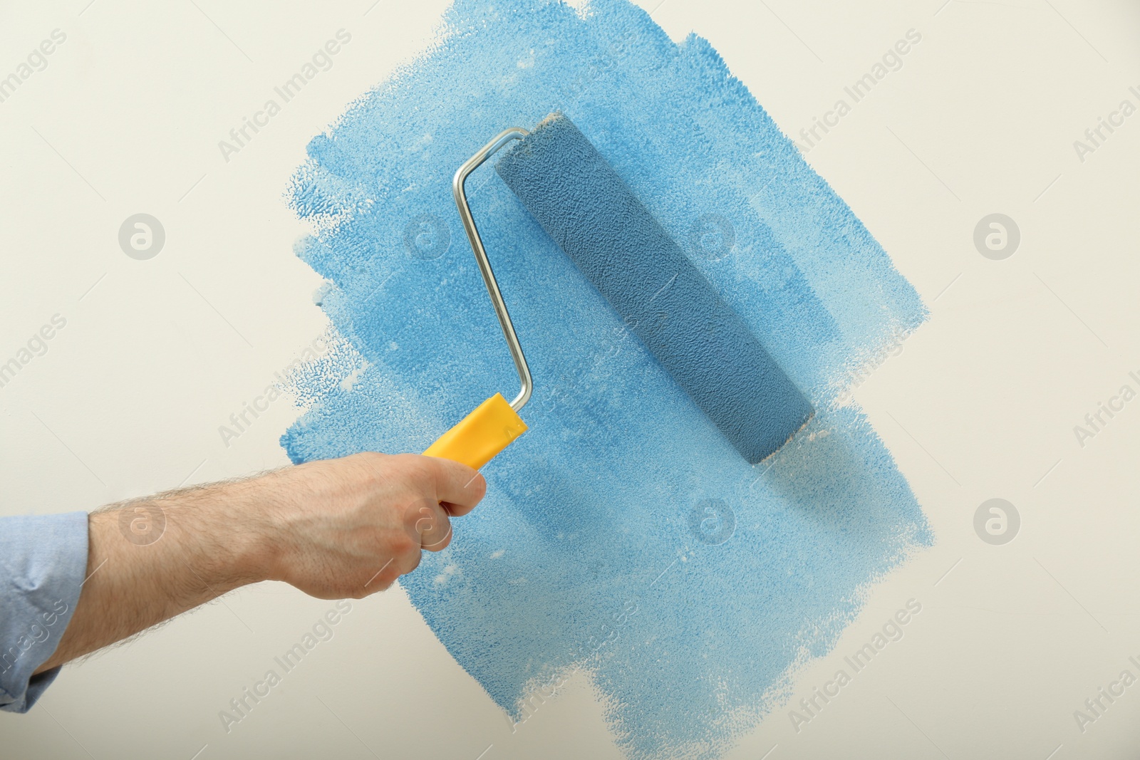 Photo of Man applying light blue paint with roller brush on white wall, closeup