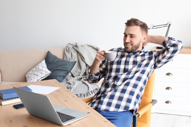 Photo of Young man drinking coffee while working with laptop at desk. Home office