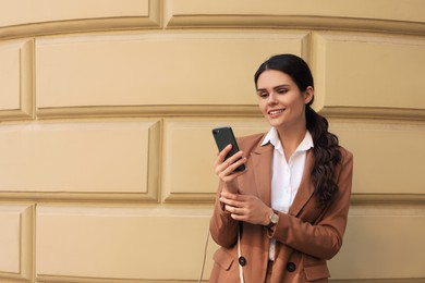 Photo of Young beautiful woman in stylish suit using smartphone near pale yellow wall outdoors