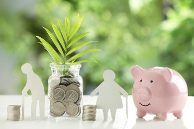 Photo of Pension savings. Figures of elderly people, piggy bank, jar with coins and twig on white table against blurred green background