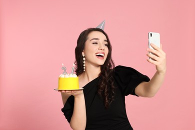 Photo of Coming of age party - 21st birthday. Smiling woman holding delicious cake with number shaped candles and taking selfie against pink background