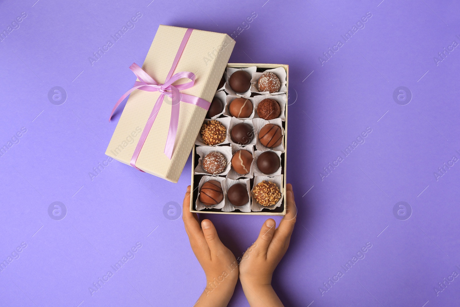 Photo of Child with box of delicious chocolate candies on light purple background, top view