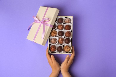 Photo of Child with box of delicious chocolate candies on light purple background, top view