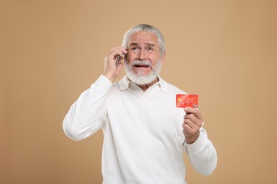 Photo of Stressful senior man with credit card talking on smartphone against beige background. Be careful - fraud
