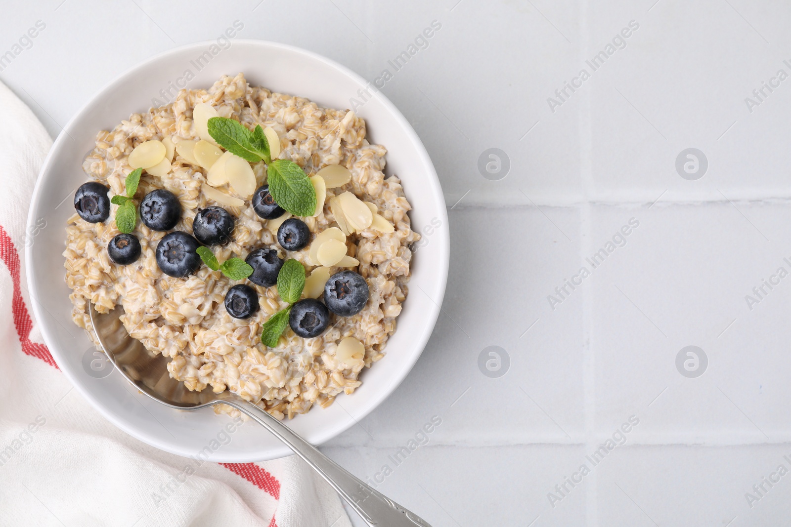 Photo of Tasty oatmeal with blueberries, mint and almond petals in bowl on white tiled table, top view. Space for text