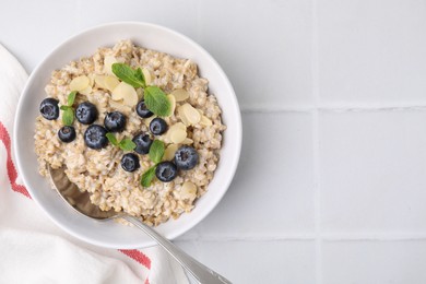 Photo of Tasty oatmeal with blueberries, mint and almond petals in bowl on white tiled table, top view. Space for text