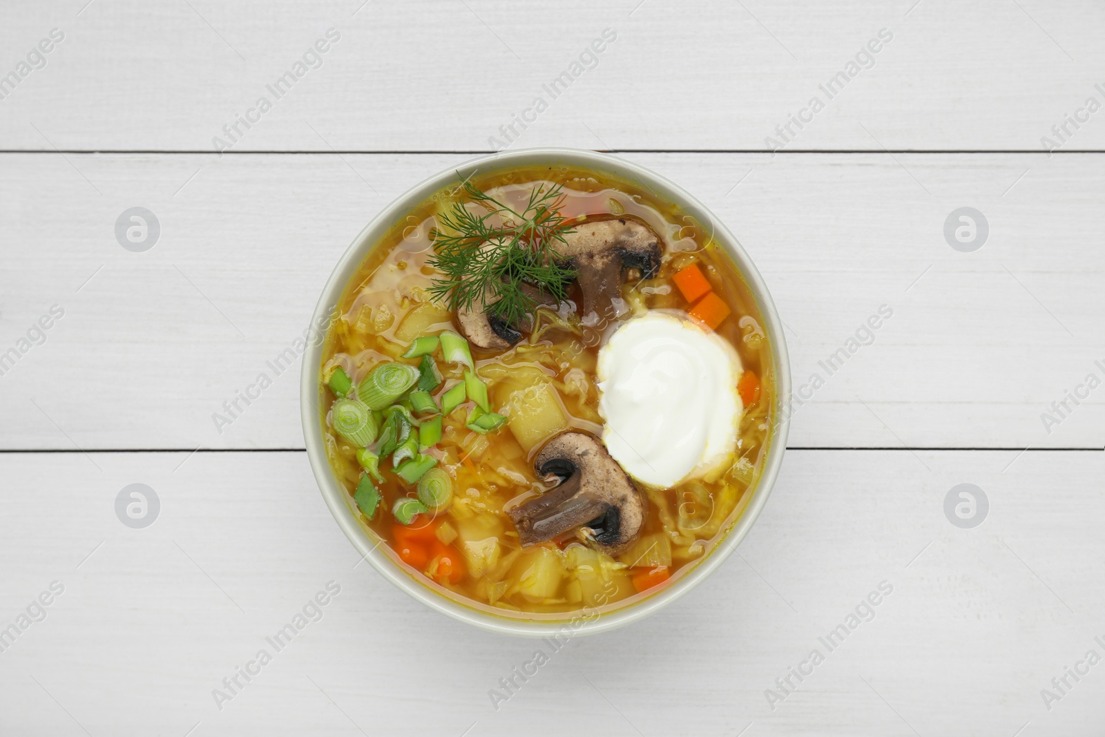 Photo of Bowl of delicious sauerkraut soup with mushrooms, green onion, dill and sour cream on white wooden table, top view