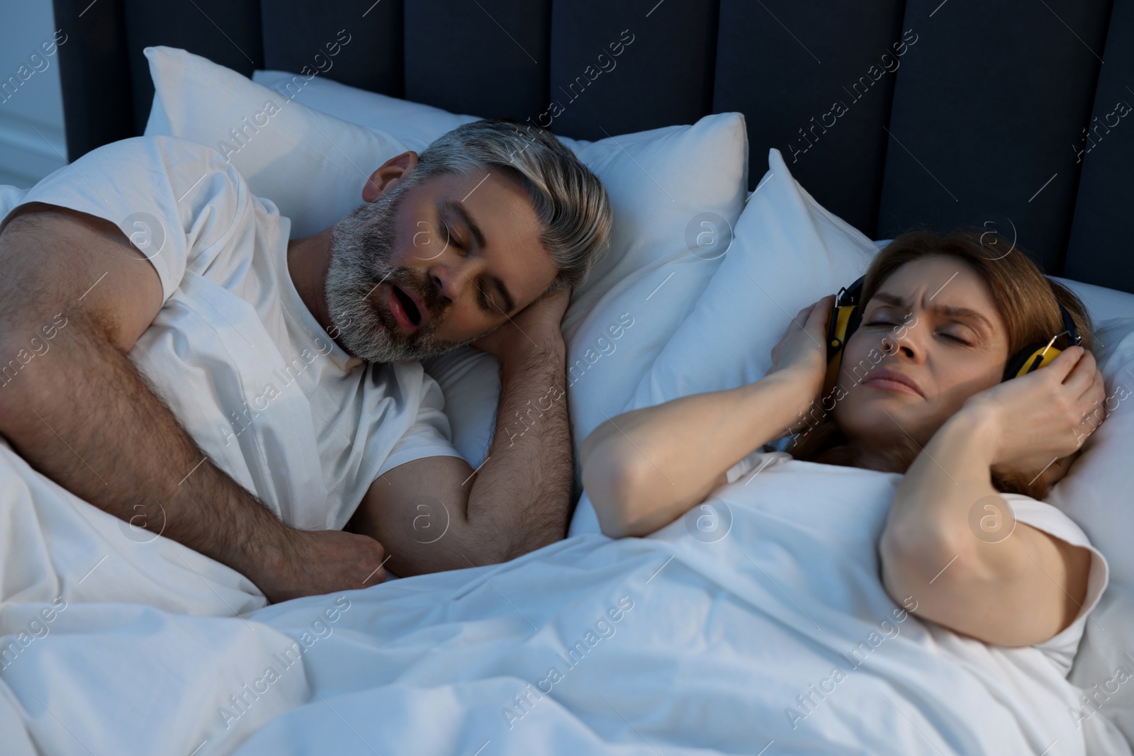 Photo of Irritated woman with headphones lying near her snoring husband in bed at home