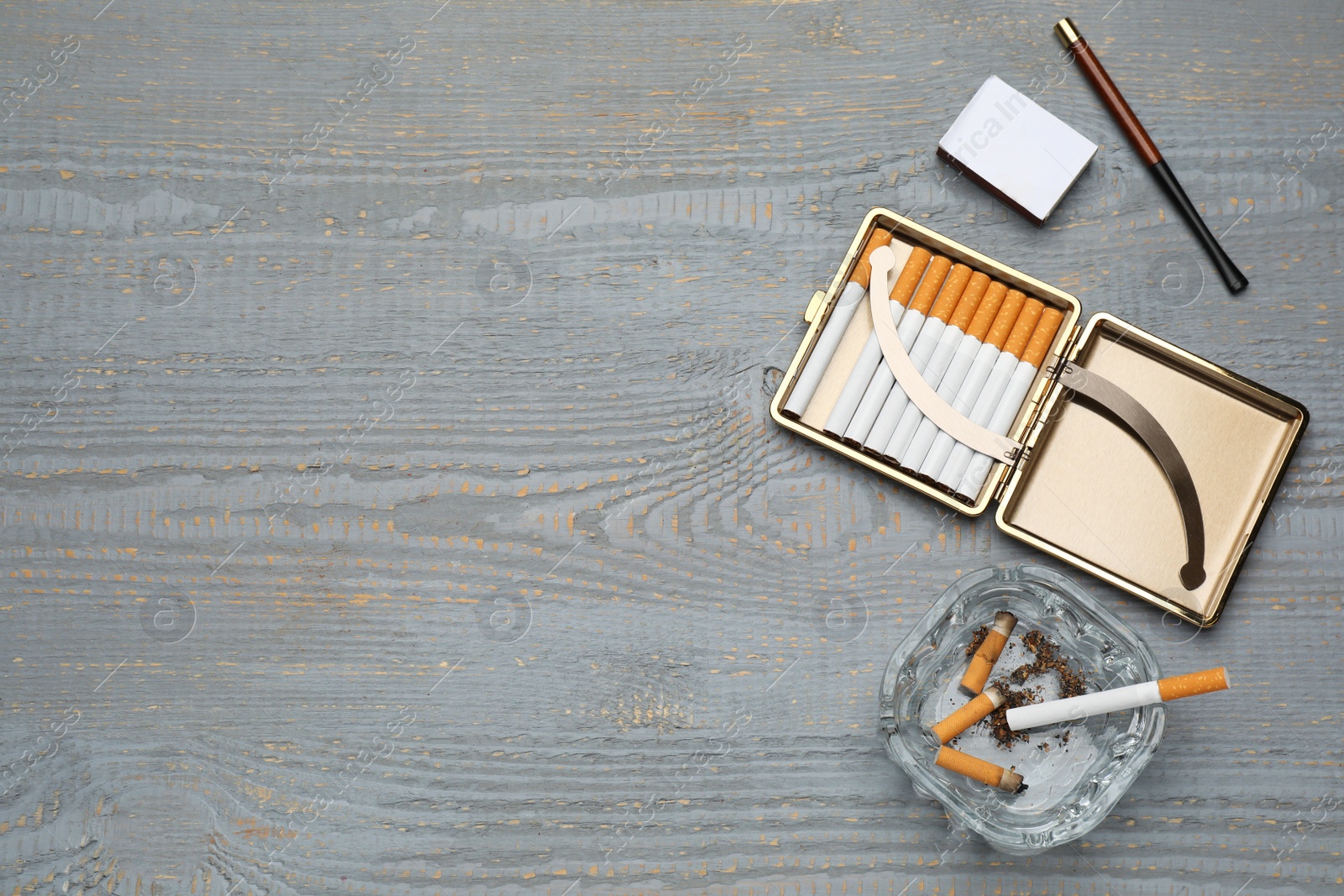 Photo of Cigarettes in case, holder, matchbox and ashtray with stubs on grey wooden table, flat lay. Space for text